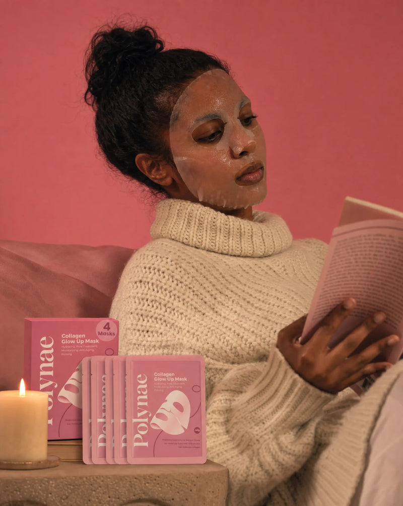Woman in a cream knit sweater relaxing with a Polynae sheet mask on her face while reading a book, candles and mask packets on a side table