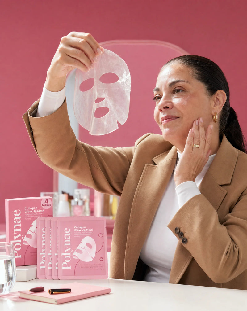 Older woman in a camel blazer holding a Polynae sheet mask up to the light and touching her cheek, multiple mask packets and boxes arranged on the table