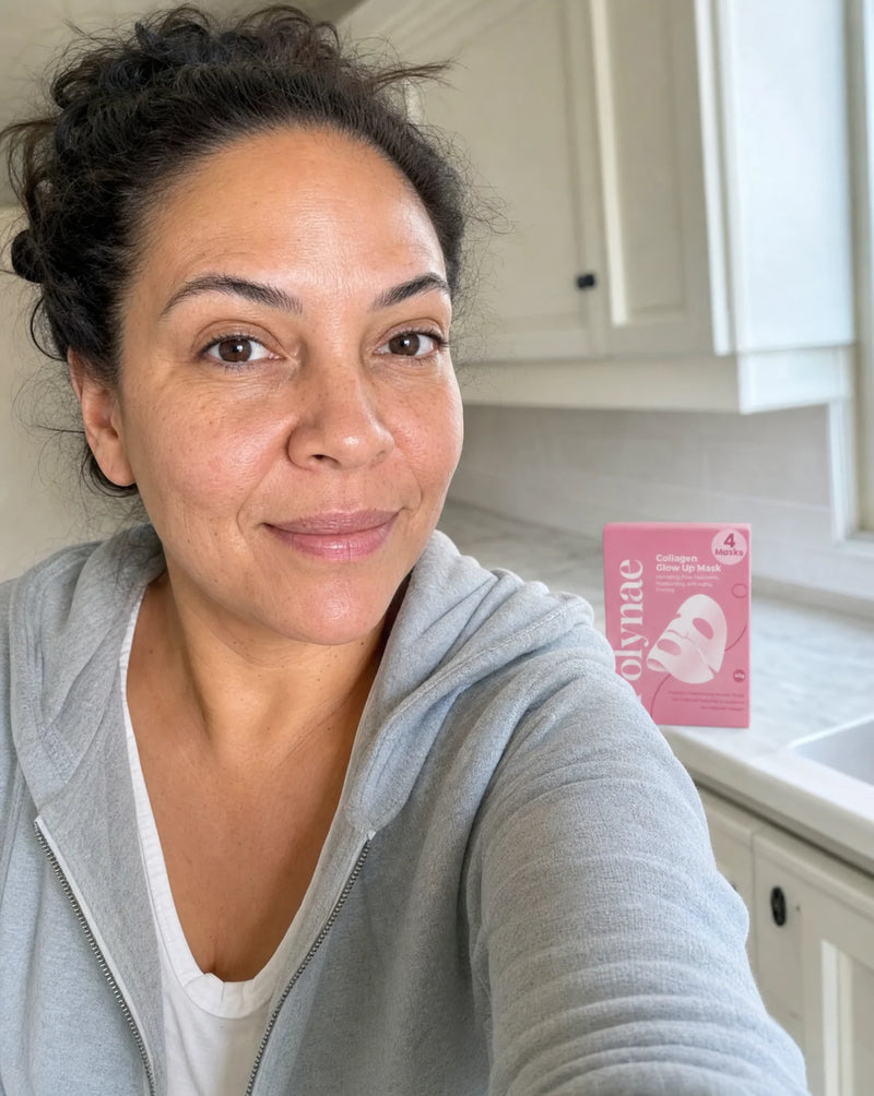 Selfie of a woman in a gray hoodie beside the Polynae Collagen Glow Up Mask box on a kitchen counter, smiling naturally