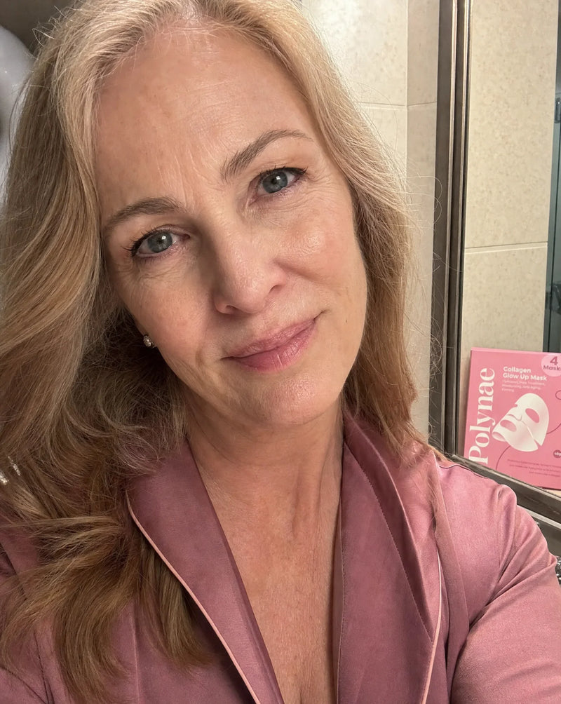 Selfie of a woman in a pink satin robe standing in a tiled bathroom with the Polynae Collagen Glow Up Mask box visible behind her