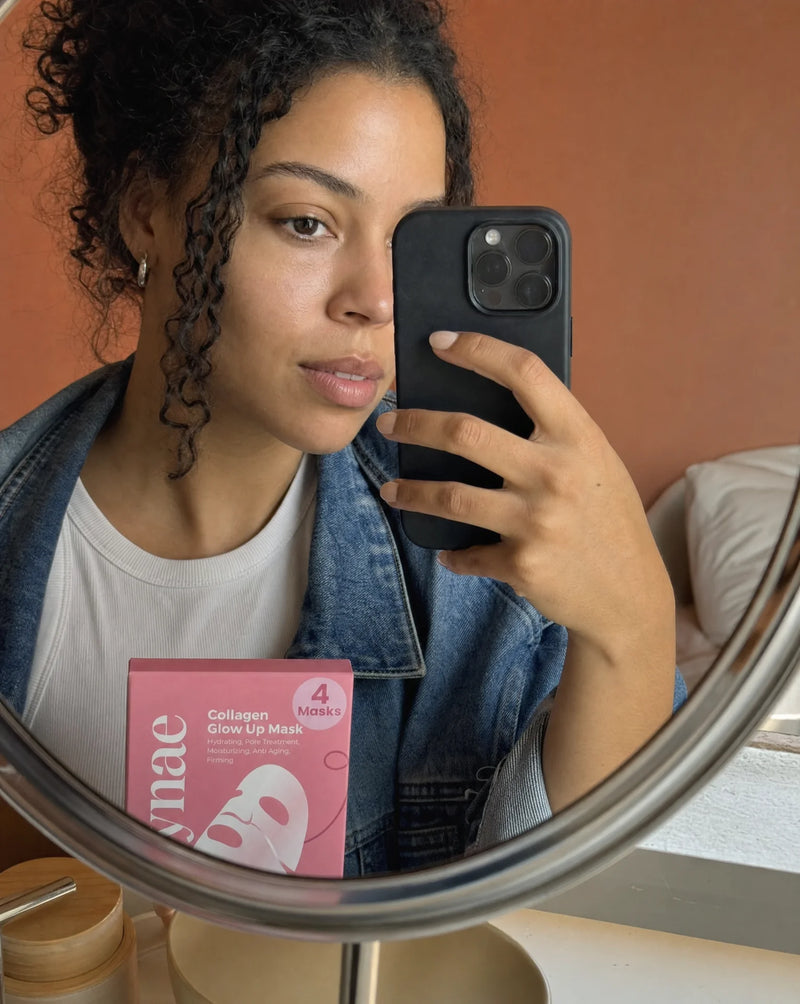 Mirror selfie of a woman in a denim jacket holding a Polynae Collagen Glow Up Mask box reflected in a gold-framed vanity mirror