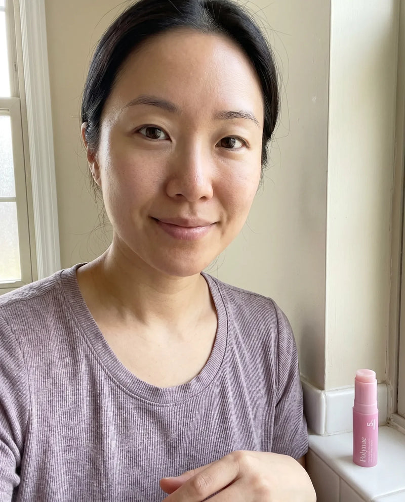 Woman with dark hair pulled back smiling softly, holding a pink Polynae lip balm stick near a window